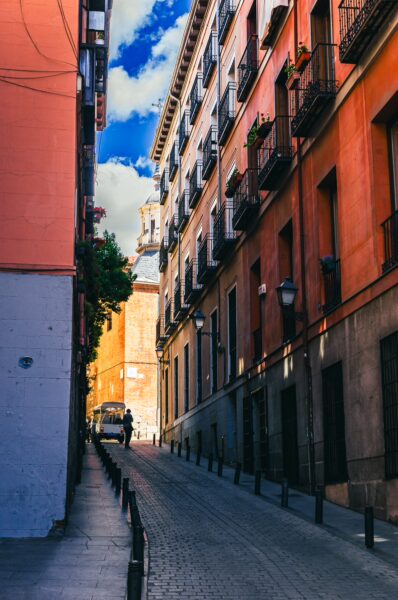 Colorful buildings line a narrow street in Madrid, Spain, with sunlight illuminating the cobblestone path and a clear blue sky above.