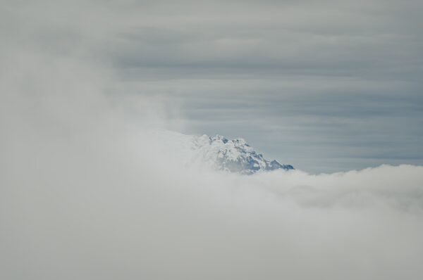 Snow-capped Mount Cotopaxi rises majestically above a sea of clouds, creating a stunning contrast with the overcast sky.