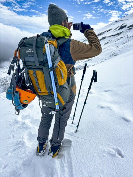 Climber wearing a bright green jacket and gray backpack takes a photo, snow-covered terrain near Chimborazo Volcano, Ecuador.