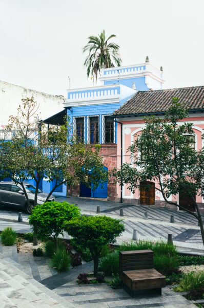 Vibrant buildings in blue and pink surround a lush garden area with low greenery at Plaza Huerto San Agustín, Quito, Ecuador.