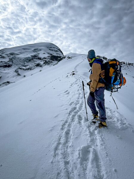 Climber ascends snowy slopes of Chimborazo Volcano in Ecuador, against a backdrop of dramatic clouds and rugged terrain.