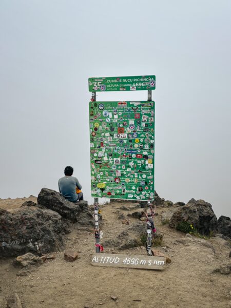 Rucu Pichincha volcano signpost surrounded by rocky terrain and a lone figure, with mist obscuring the landscape in Ecuador.