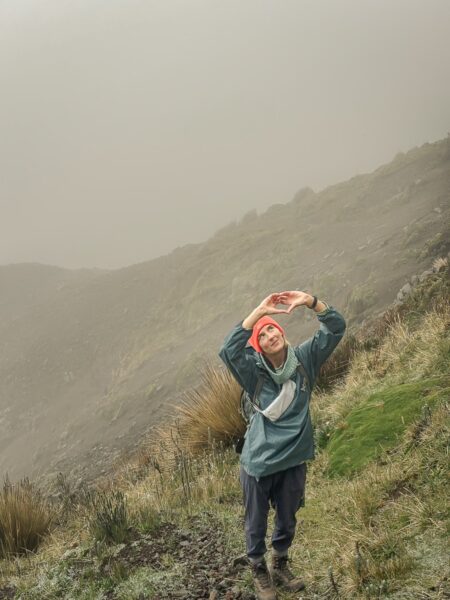A traveler with a red hat poses with arms raised, surrounded by misty grasslands and rugged terrain near Ruminahui, Ecuador.