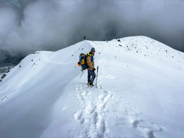 Snow-covered summit of Chimborazo Volcano, with a solitary climber navigating the ridge under a cloudy sky in Ecuador.