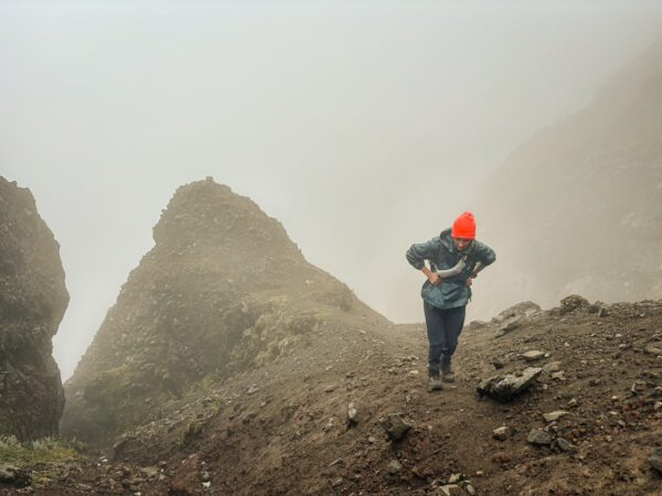 Hiker with an orange beanie navigates rocky terrain near Volcán Ruminahui, surrounded by misty mountains in Ecuador.