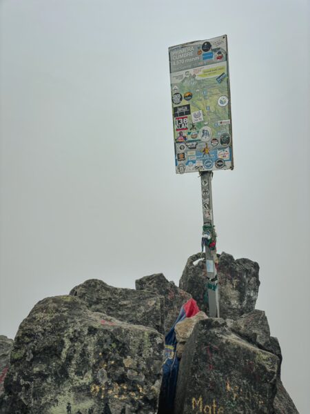 Summit marker covered in stickers atop Imbabura Volcano, surrounded by rocky terrain and a misty atmosphere in Ecuador.