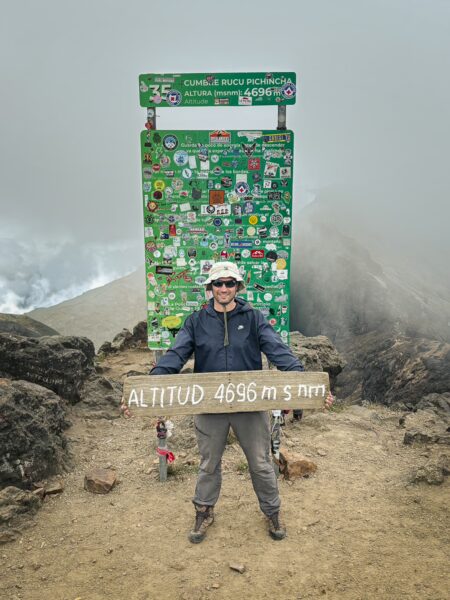 A hiker stands at the Rucu Pichincha Volcano summit sign, surrounded by rocky terrain and misty clouds in Ecuador.