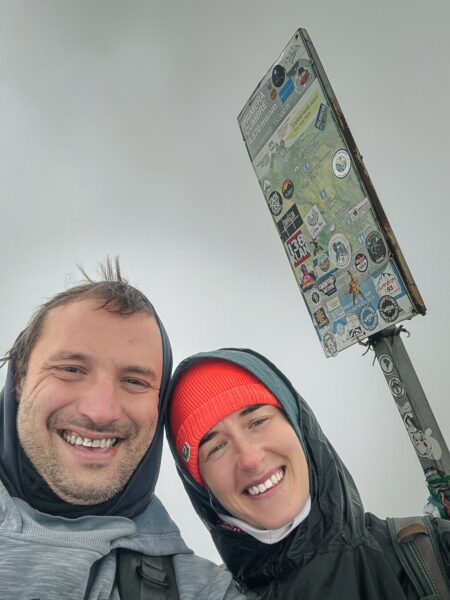 Smiling hikers at the summit of Imbabura Volcano, with a sticker-covered signpost and cloudy skies in Ecuador.