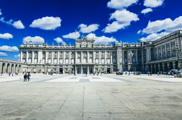 Elegant view of Palacio Real under a bright blue sky, with fluffy clouds and visitors exploring the grand courtyard in Madrid, Spain.