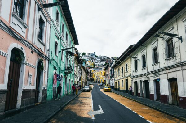 Colorful colonial buildings line the narrow street in Quito, Ecuador, with a cloudy sky overhead and a glimpse of the hillside in the background.