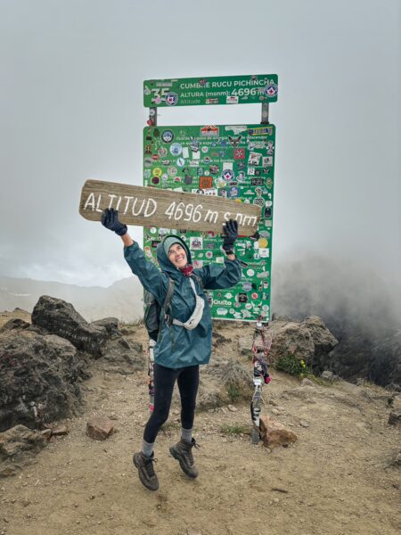 Rucu Pichincha Volcano's summit sign shows altitude of 4695 m, with a climber celebrating amidst rocky terrain and misty surroundings.
