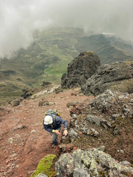 Climber navigating rocky terrain on Ruminahui Volcano, shrouded in mist, with green hills visible in the background, Ecuador.