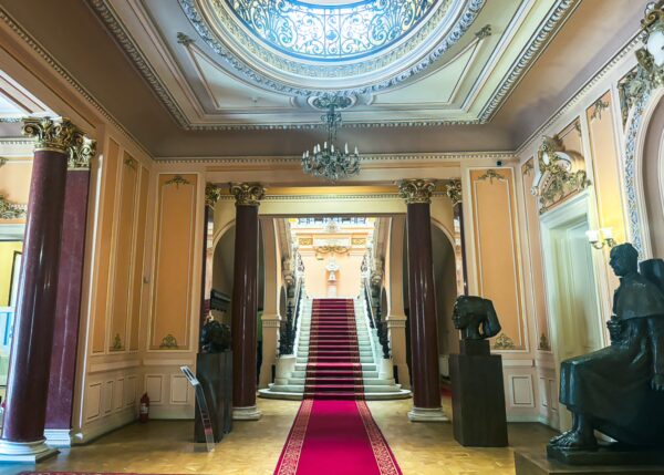 Elegant interior of the Art Museum in Craiova, Romania, featuring a grand staircase, red carpet, and classic sculptures.