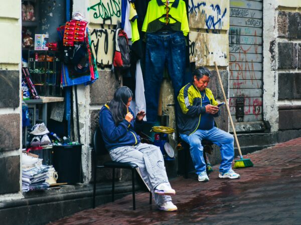 Vibrant street scene in Guayaquil features two men sitting near a shop, surrounded by colorful clothing and graffiti.