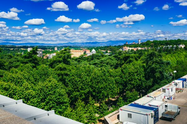 Lush green trees fill the foreground, with a vibrant sky and distant mountains visible from the Royal Palace ledge viewpoint in Madrid.