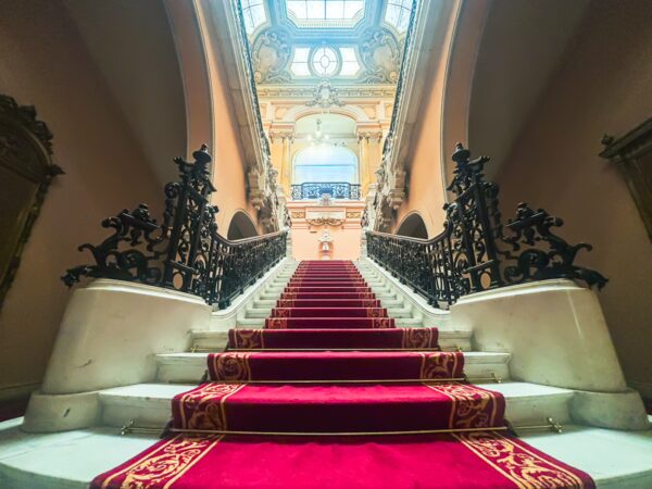 Red carpeted staircase with ornate railings leads to a beautifully illuminated upper level in the Art Museum, Craiova, Romania.