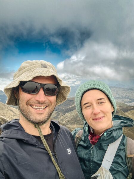 Smiling hikers wearing hats and jackets pose against a backdrop of cloudy skies on Rucu Pichincha Volcano, Ecuador.
