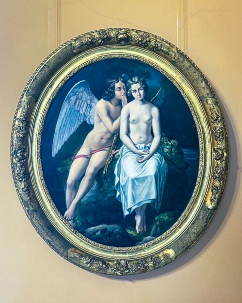 Colorful painting in ornate frame depicting a winged figure whispering to a seated youth at the art museum in Craiova, Romania.