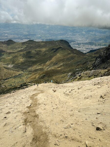 Sand-covered trail leads down Pichincha Volcano, showcasing lush green hills and a distant cityscape in Ecuador.