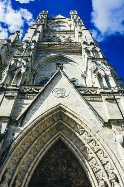 Basilica del Voto Nacional's intricate stone façade reaches skyward under a blue sky with fluffy clouds in Quito, Ecuador.