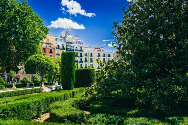 Lush hedges and manicured gardens frame vibrant buildings under a bright blue sky at Plaza de Oriente, Madrid.