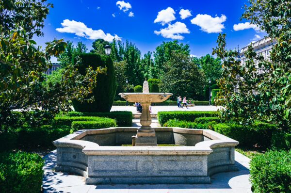 A stone fountain surrounded by lush greenery and well-trimmed hedges at Plaza de Oriente, Madrid, Spain, under a bright blue sky.