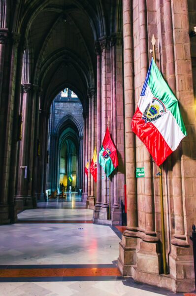 Stained glass and stone arches frame colorful flags inside Basilica del Voto Nacional in Quito, Ecuador.