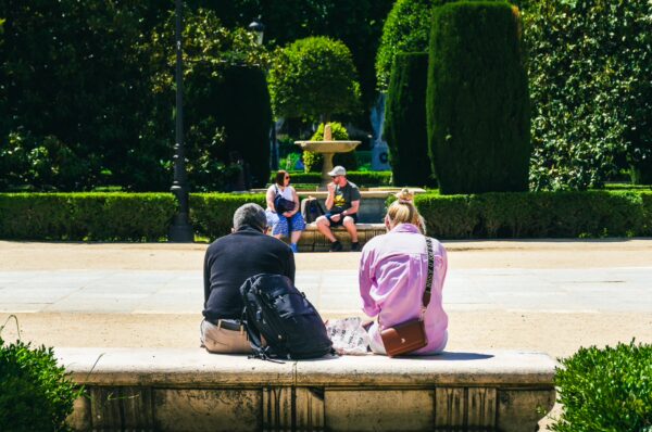 Relaxing visitors sit on a stone bench in Plaza de Oriente, Madrid, surrounded by neatly trimmed hedges and a fountain.