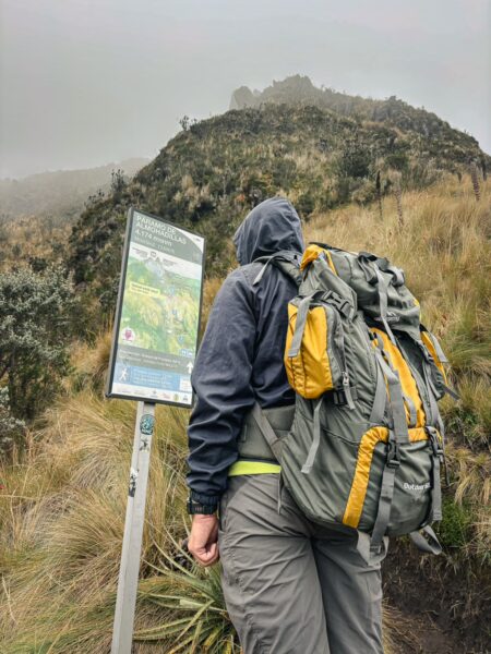 Hiker in a gray and yellow backpack studies a trail map near Imbabura Volcano, surrounded by lush green vegetation in Ecuador.