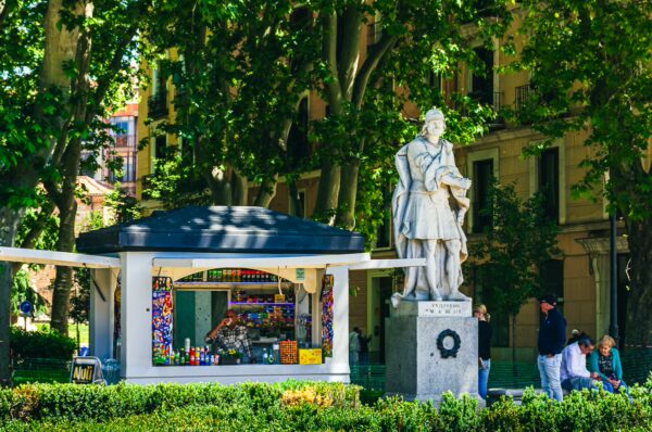 Statue and kiosk in Plaza de Oriente, Madrid, surrounded by lush greenery and vibrant city architecture.
