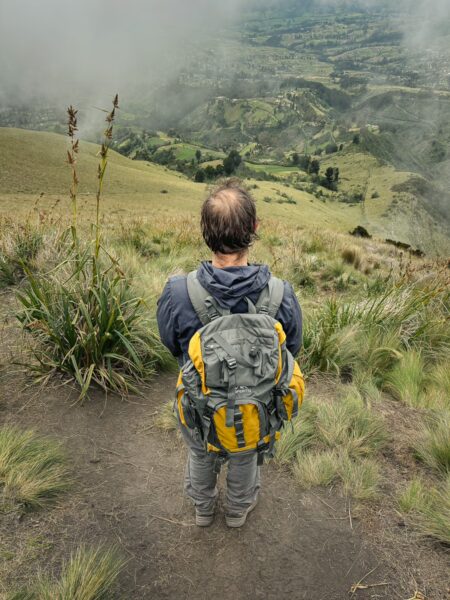 Hiker in a gray jacket with a yellow backpack stands on a grassy trail, overlooking the lush landscape of Imbabura Volcano, Ecuador.