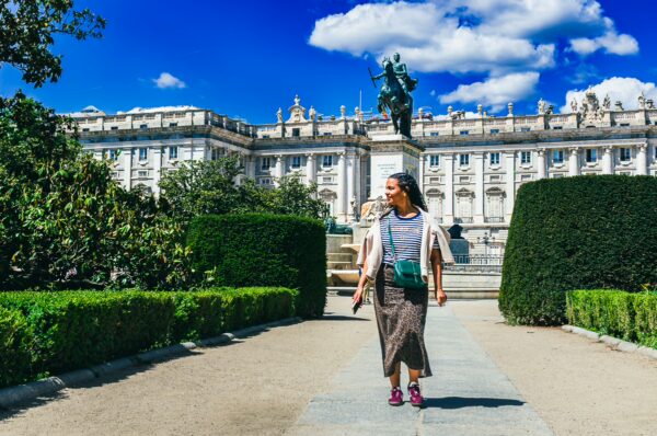 A woman strolls through lush greenery in Plaza de Oriente, with the Royal Palace of Madrid and blue skies in the background.