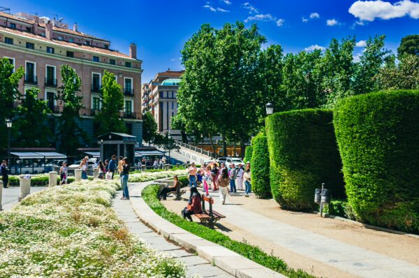 Lush greenery and blooming flowers surround visitors enjoying the vibrant atmosphere of Plaza de Oriente in Madrid, Spain.