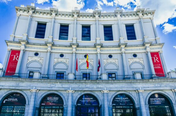 Teatro Real features elegant white architecture with decorative columns, banners, and a blue sky above in Madrid, Spain.