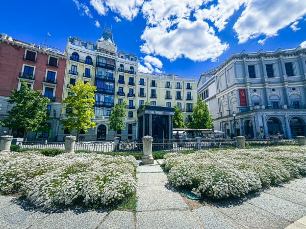 Vibrant flowers in full bloom contrast with the historic buildings of Plaza de Oriente, framed by a bright blue sky in Madrid.