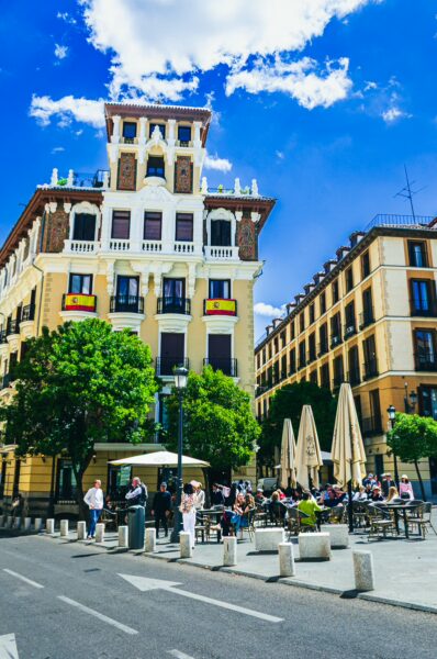 Vibrant plaza in Madrid features café seating, lush green trees, and ornate buildings under a bright blue sky.