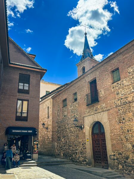 Charming street scene in Madrid, Spain, featuring historic brick buildings, a clock tower, and a bright blue sky.