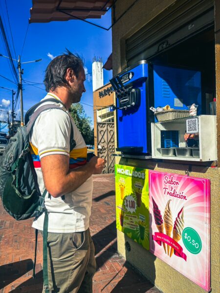 A traveler using a blue vending machine in Quito, Ecuador, with colorful posters nearby and a clear sky overhead.