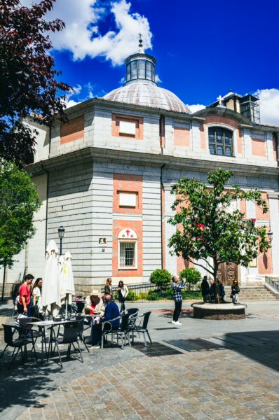 Visitors enjoy outdoor seating near a distinctive pink and gray building under a blue sky in Madrid, Spain.