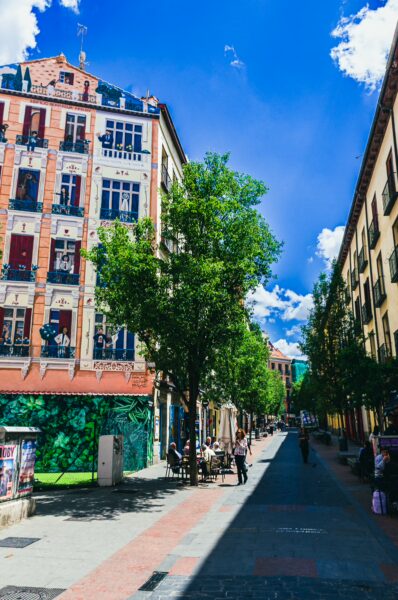 Sunny street scene in Madrid, Spain, featuring vibrant buildings, greenery, and pedestrians enjoying a lively atmosphere.