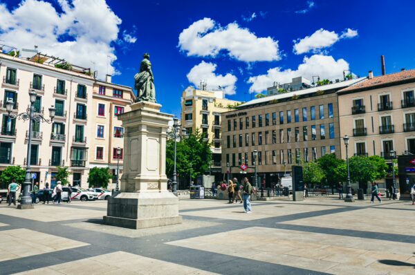 Statue surrounded by sunlit buildings and lively pedestrians in vibrant Plaza de España, Madrid, Spain. Bright blue sky with clouds.