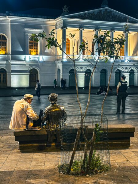 Two figures sit on a bench, with a bronze statue nearby, framed by the illuminated Plaza del Teatro in Quito, Ecuador.