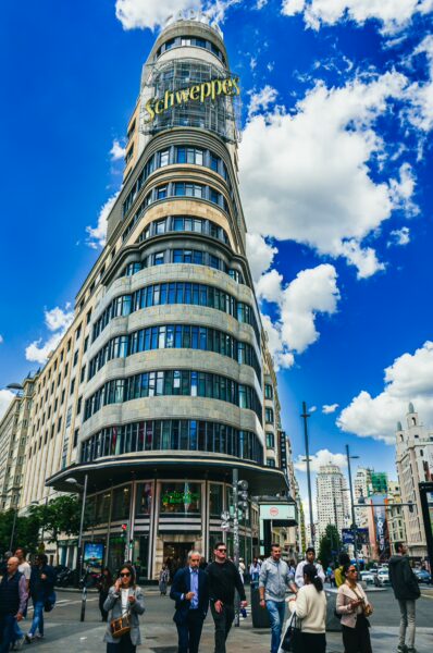 Schweppes sign prominently displayed on the iconic Metropolis building in Madrid, with a bustling street scene beneath a bright blue sky.