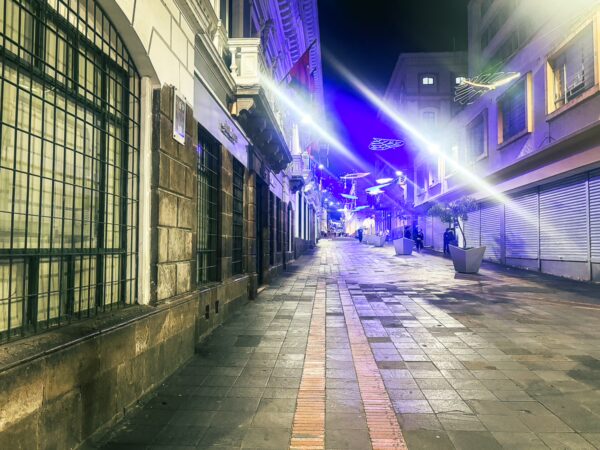 Vibrant night scene in Quito, Ecuador, featuring a cobblestone street illuminated by colorful lights and empty shops.