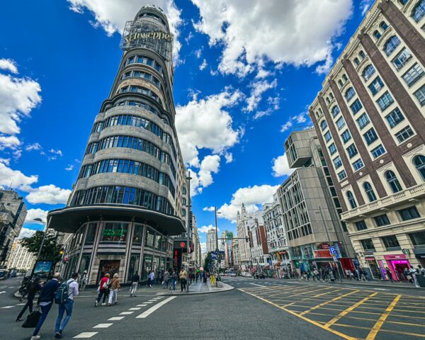A vibrant street view in Madrid features the iconic Schweppes building against a backdrop of blue skies and scattered clouds.