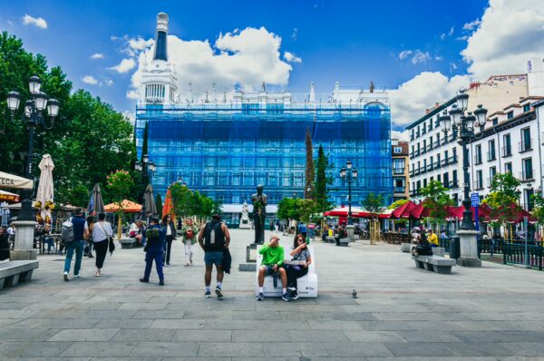 Vibrant plaza de Santa Ana in Madrid, featuring lively cafes, trees, and a blue sky with scattered clouds.