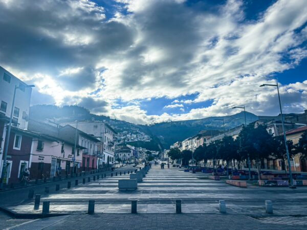 Sunlight breaks through clouds over a wide plaza in Quito, Ecuador, lined with trees and historical buildings in soft pastel colors.