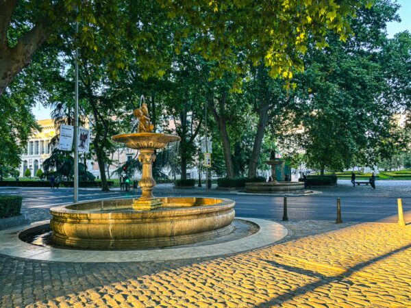 Tranquil fountain surrounded by lush trees along Paseo del Prado in Madrid, Spain, reflecting soft evening light.