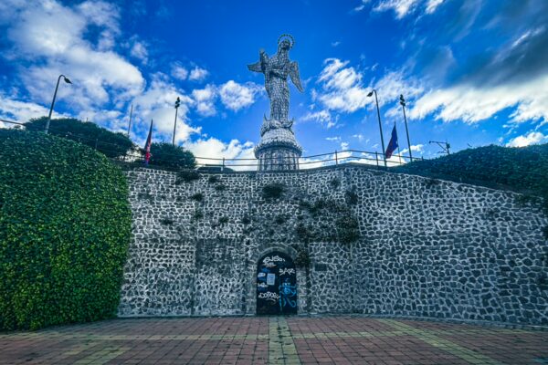 El Panecillo statue stands majestically atop a stone wall, surrounded by lush greenery and a vibrant sky in Quito, Ecuador.