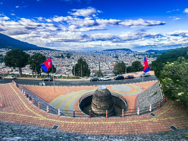 Scenic view over Quito, Ecuador, featuring vibrant flags, a circular stone structure, and rolling hills under a bright blue sky.